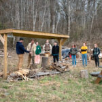 Seven people watch as two men prepare to cut a wood chunk outside.