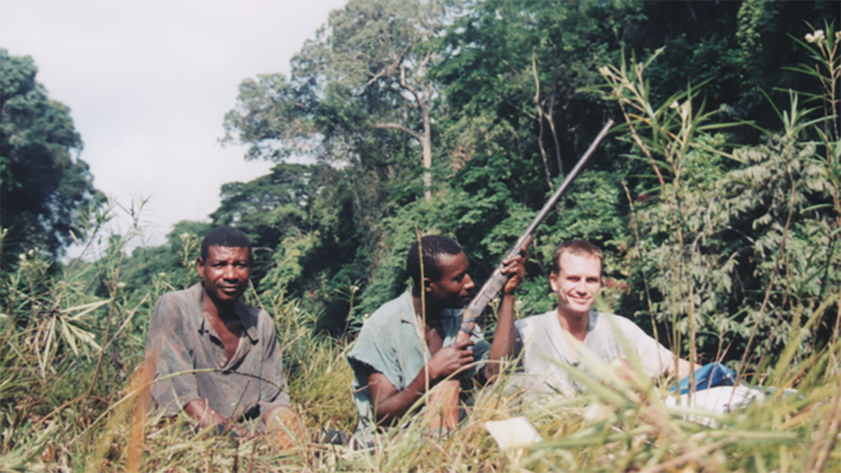 Three men sit in the grass, and one holds a gun.