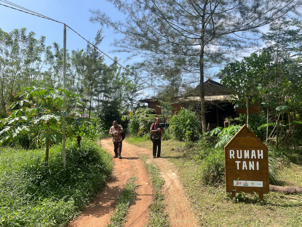 Two people stand on dirt path near building and sign with grass and trees.