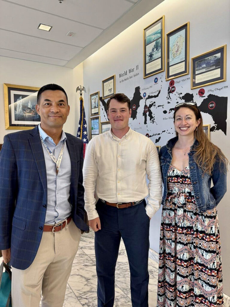 Three people smile and stand in the corner of a room next to a all covered in a map and pictures.
