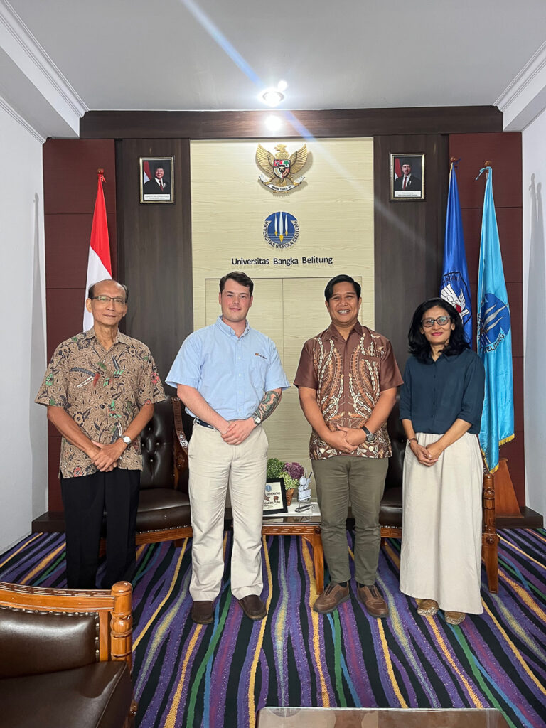 Four people stand and smiles in front of the University of Bangka Belitung seal and flags.