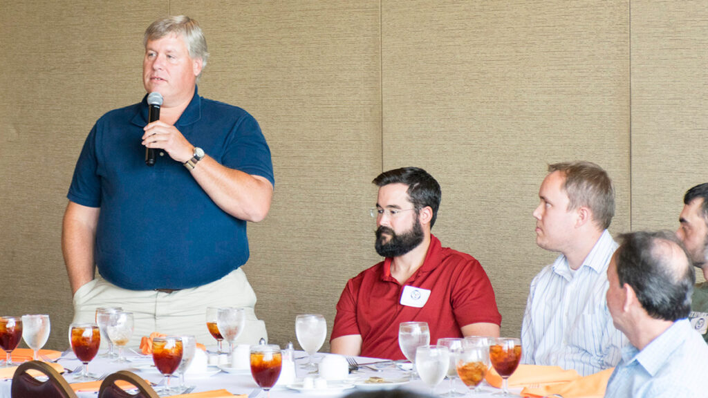 One person stands with a microphone, and four other people sit at a table.