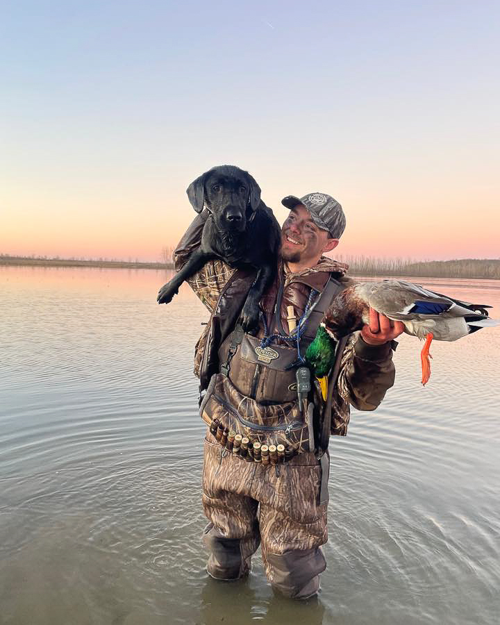 A person holds a dog and a dead duck in water.