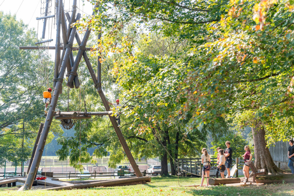 A person pauses on a climbing tower while others watch.