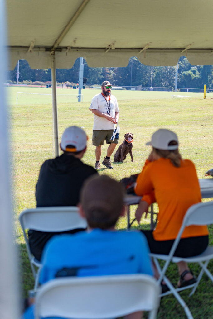 A person stands with a dog with people watching under a tent.