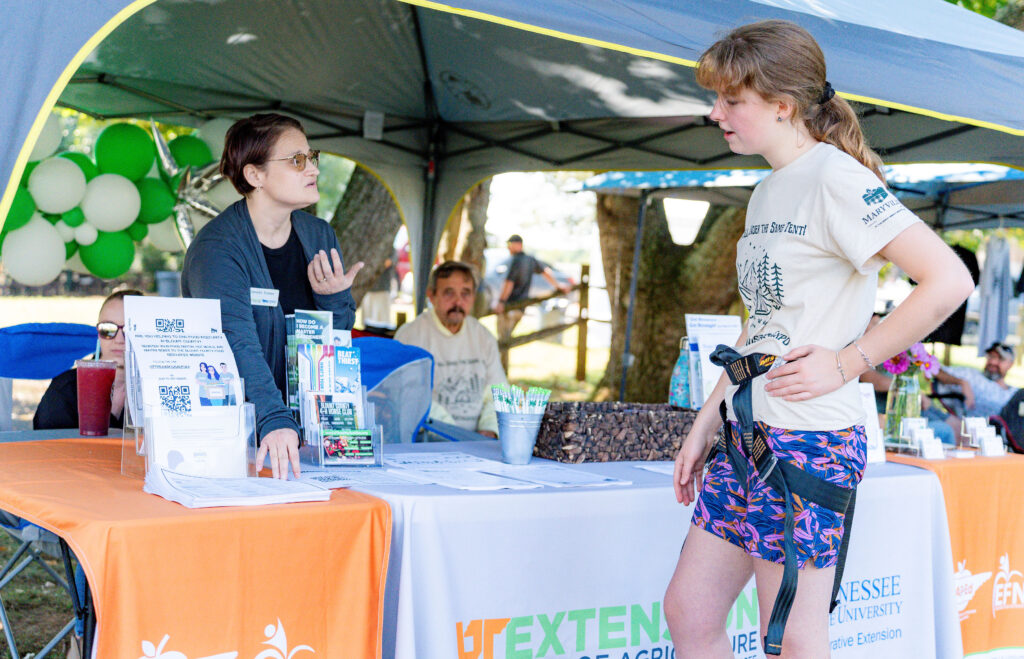 Two people talk near a table and tent with two others sitting nearby.