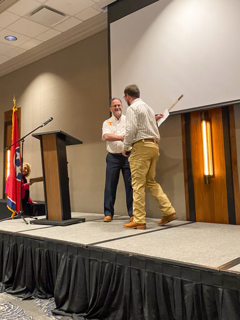 Two people shake hands on a stage with another person watching from the floor.