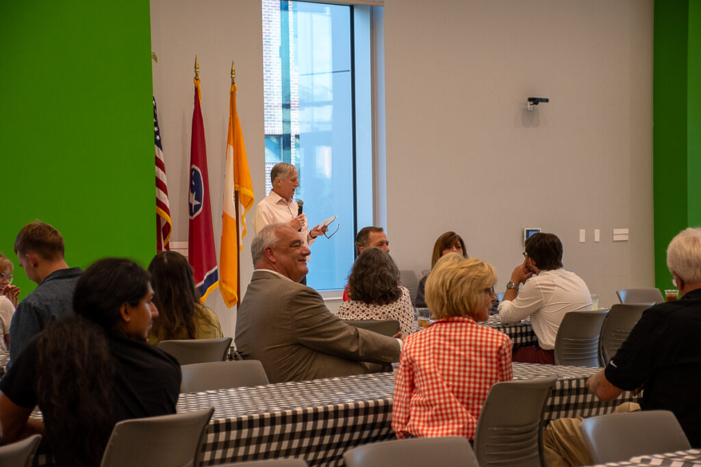 A man talks into a microphone in front of people sitting at tables in a building on the UT Institute of Agriculture campus.