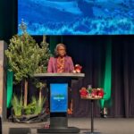 A woman stands at a podium on a stage with a projector screen behind her.