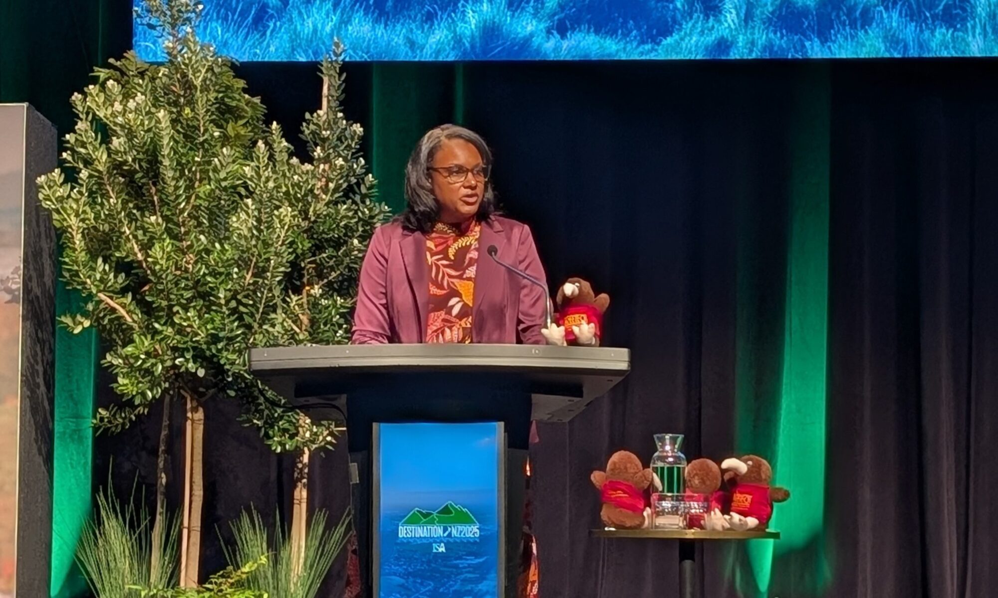 A woman stands at a podium on a stage with a projector screen behind her.