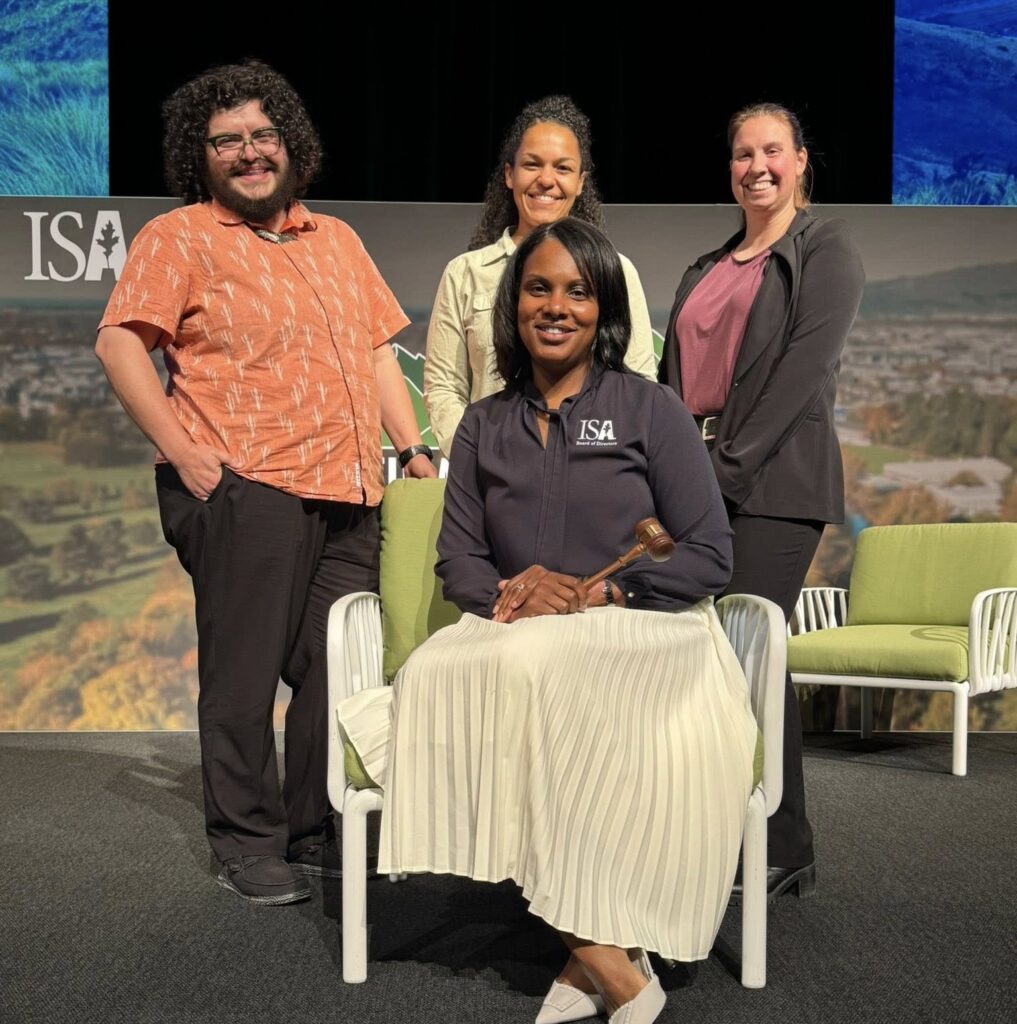 Three people stand and one person sits in a chair on a stage.