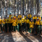 Twenty-two people wearing fire protection gear stand with equipment in forest plot at the UT Arboretum.