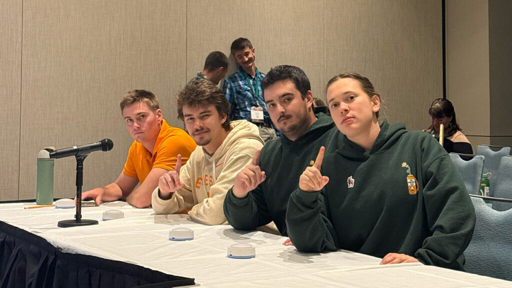 Four people sit at a table with buzzers and two people stand behind them at the Society of American Foresters annual quiz bowl.