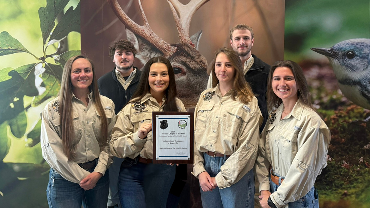 Six people stand in front of mural of deer, bird, and leaves with a plaque.