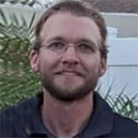 Person smiles in front of bush and white fence.