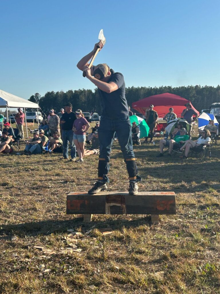 A person swings an ax while standing on a log with people behind him.