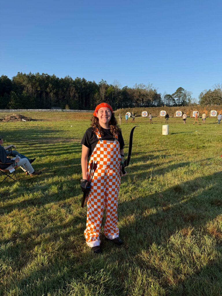 A person in checkered orange and white overalls holds a bow and arrow with targets in the background in a field.
