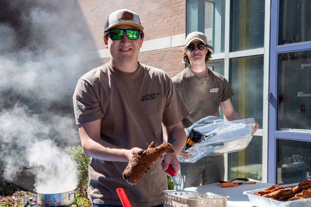 One person holds a fried fish, and another person holds an aluminum pan outside a building on the UT Institute of Agriculture campus.