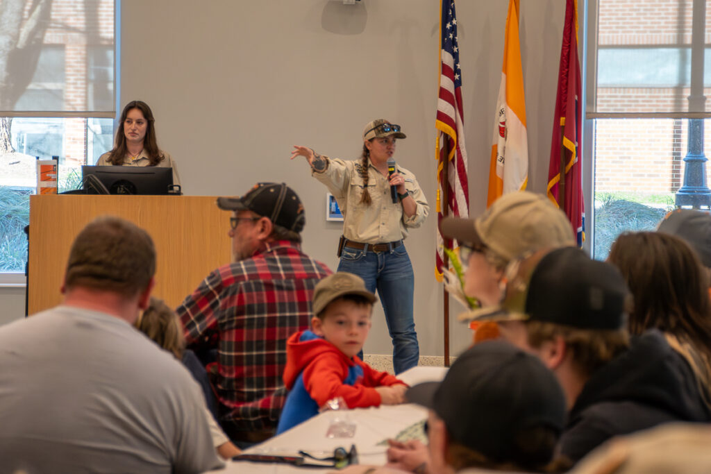 A person holds a microphone and gestures in a room with other people in a building on the UT Institute of Agriculture campus.