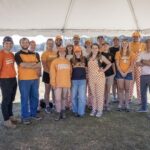 Nineteen people stand together under a tent outside.