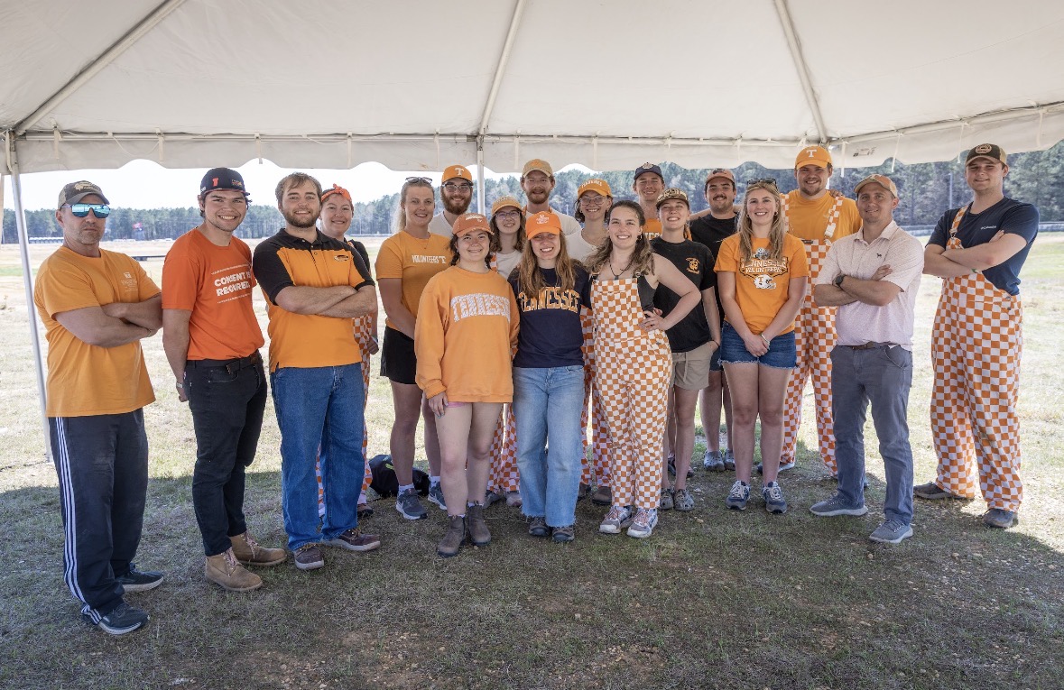 Nineteen people stand together under a tent outside.