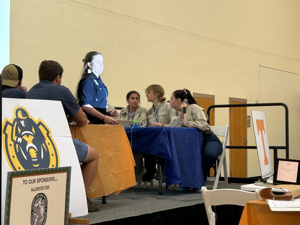 Four people sit at tables on a podium while a fifth person stands at the University of Florida.