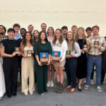 Twenty people stand in a group with seven of them holding plagues.