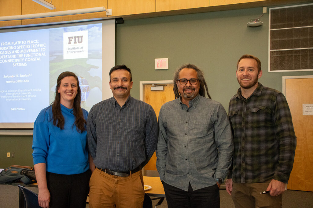 Four people stand in a room with a projector screen behind them.