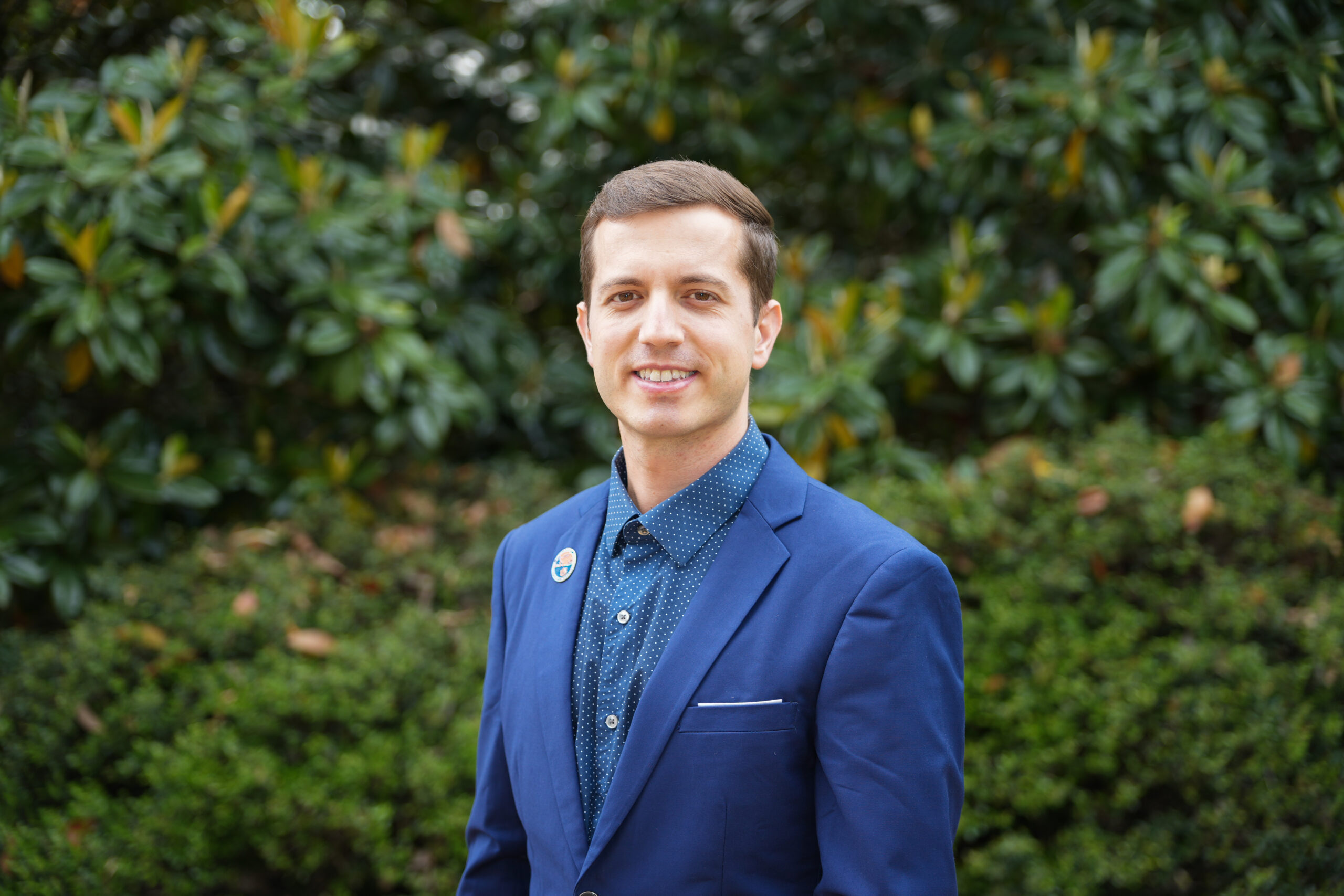 Person wearing button up and jacket stands in front of tree.