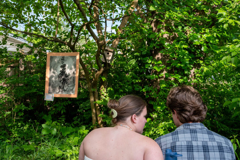 A drawing hangs from a tree with two people sitting nearby.