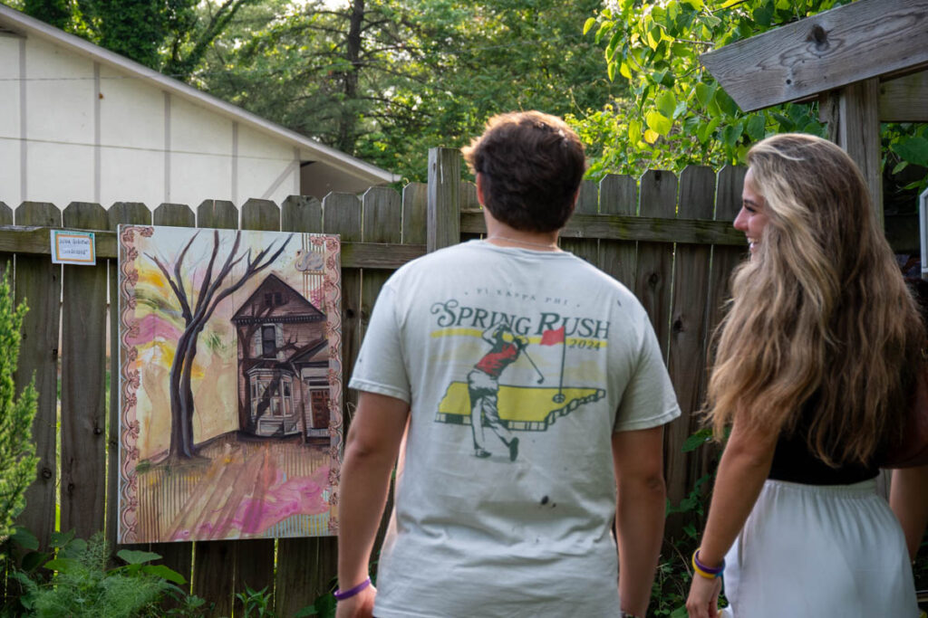 Two people look at a painting hung on a fence.