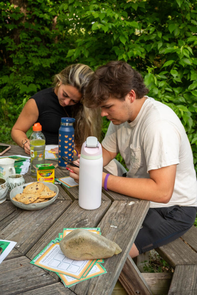 Two people draw on paper on a table with water bottles and cookies.