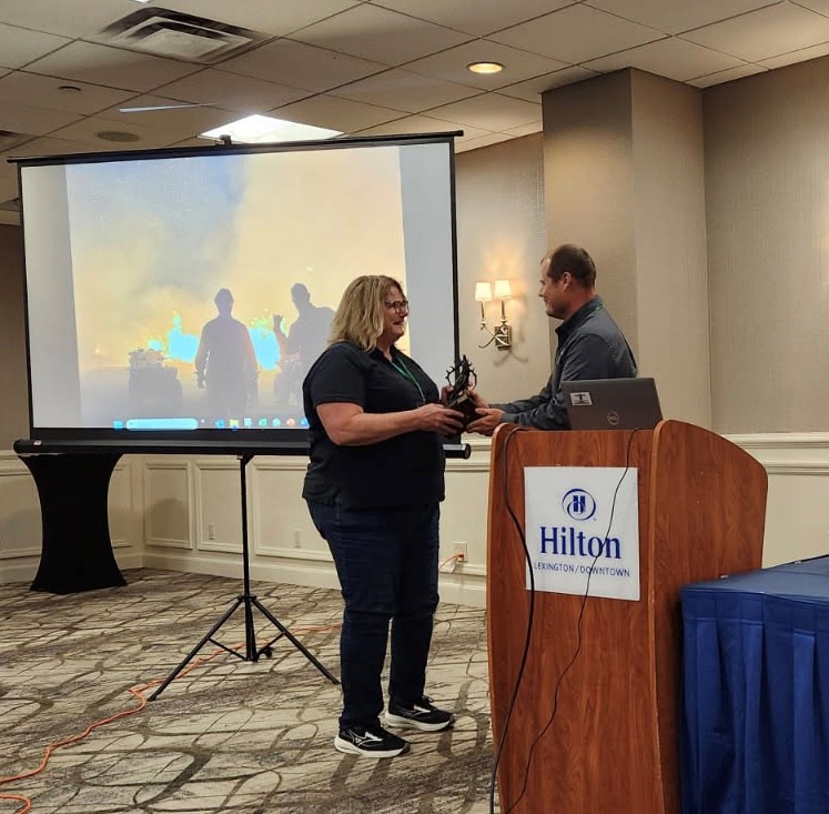 A person presents another person with an award by a podium and projector screen in a Hilton hotel in Lexington, Ky.