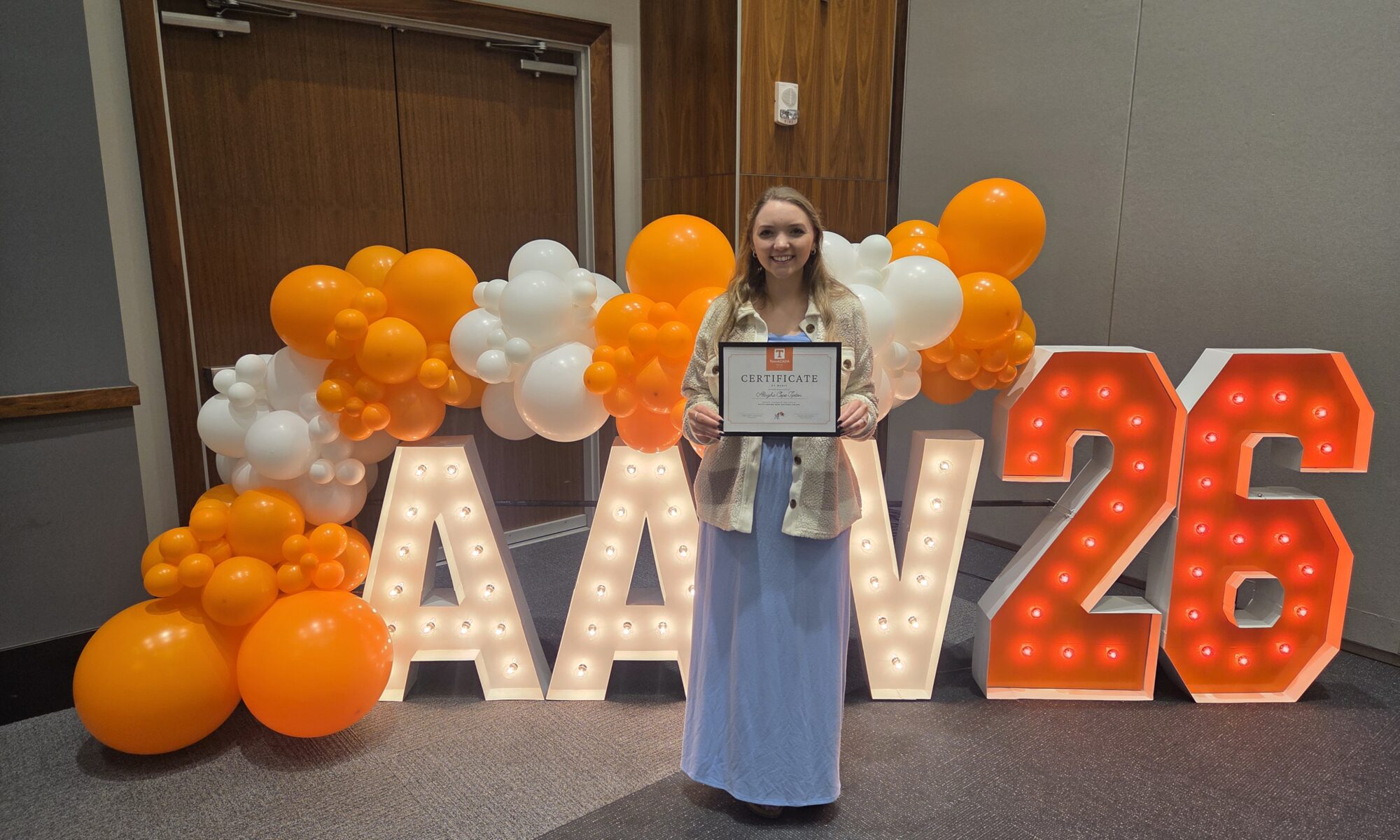 A person holds a certificate in front of a lit up sign and balloons.