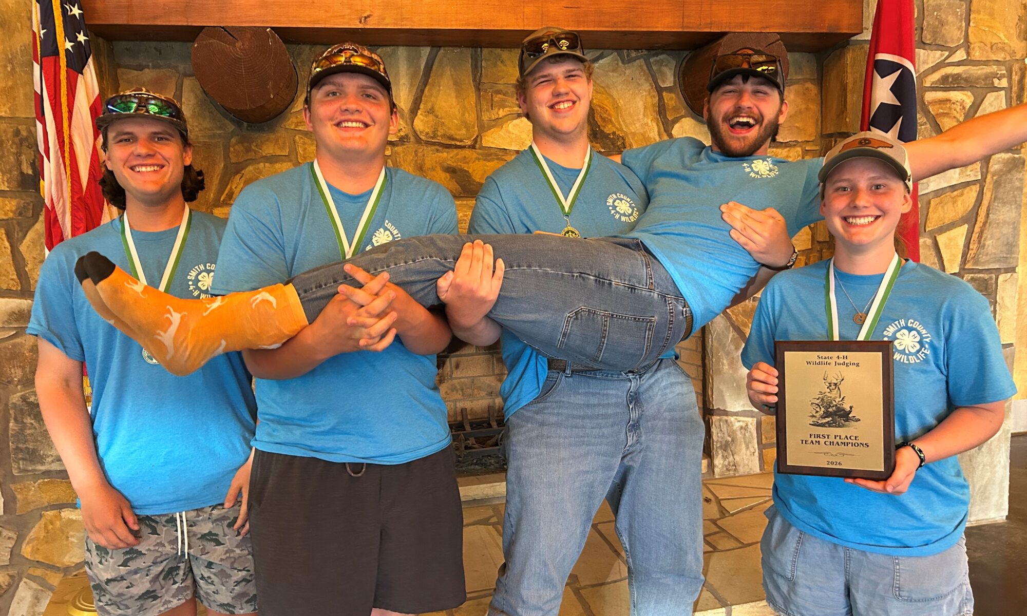 Three people hold a person with another standing beside them with a plaque in front of a fireplace.