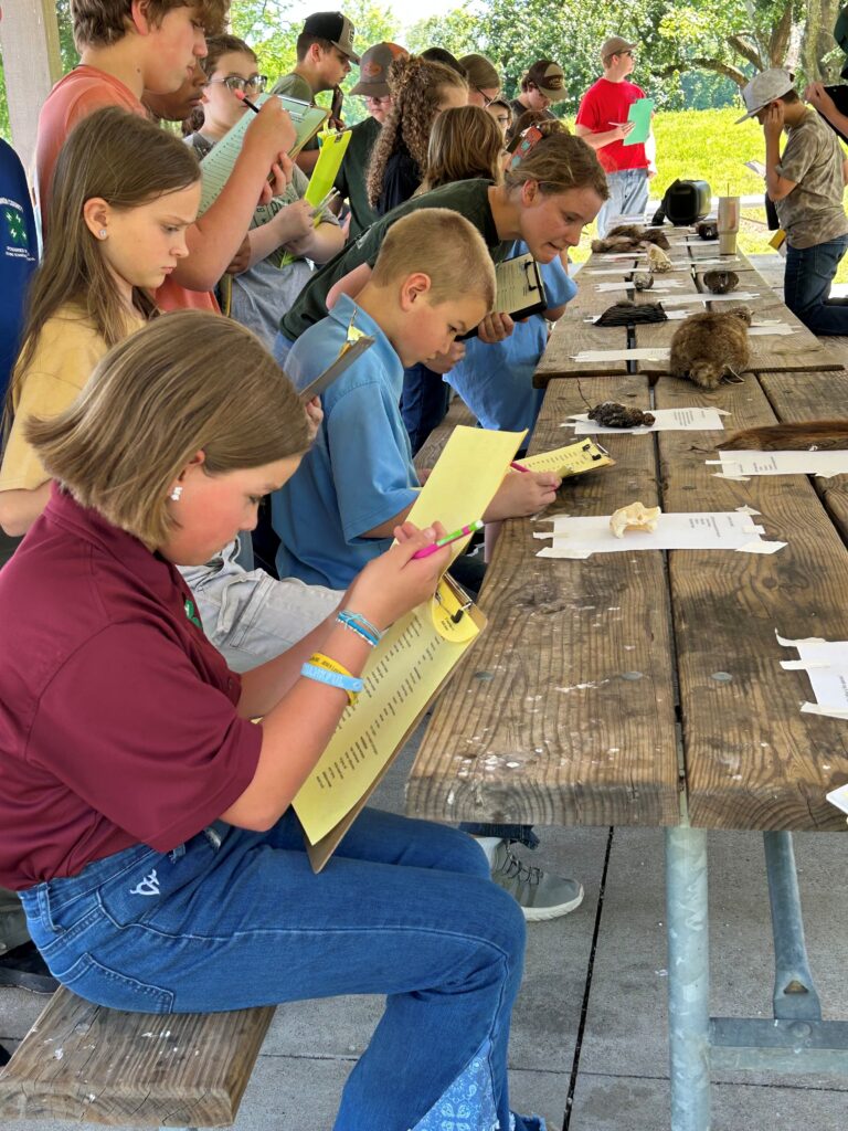 Students sit or stand at a table with notepads and pencils.