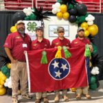 Four people hold the Tennessee flag in front of a 4-H sign with yellow and black balloons.