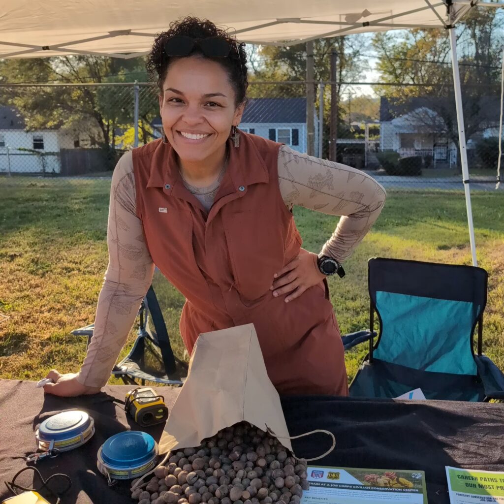 Person stands under pop up tent next to camp chair with a hand on a table next to a bag of acorns and tree measuring tools.