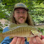 Person wearing glasses and hat holds fish near stream.