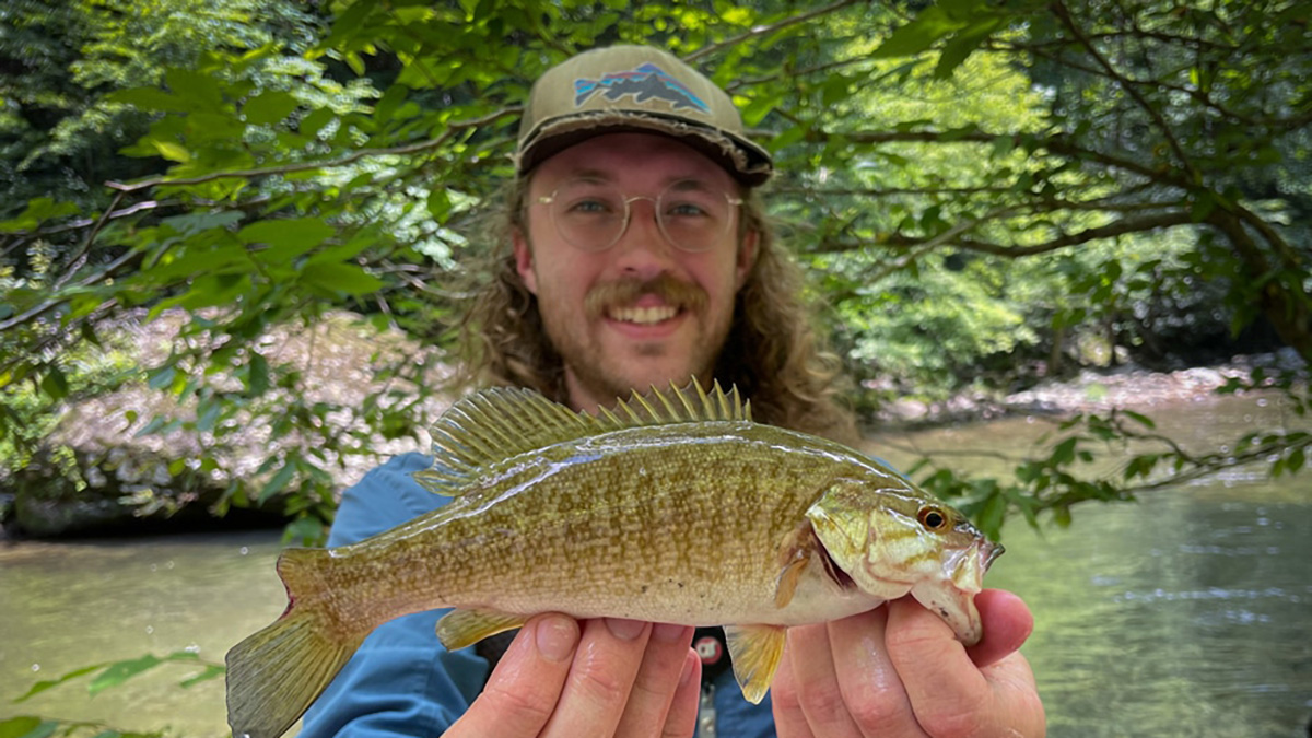 Person wearing glasses and hat holds fish near stream.