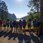 Twelve people stand next to a tree in front of a lake at the Philmont Scout Ranch in New Mexico.