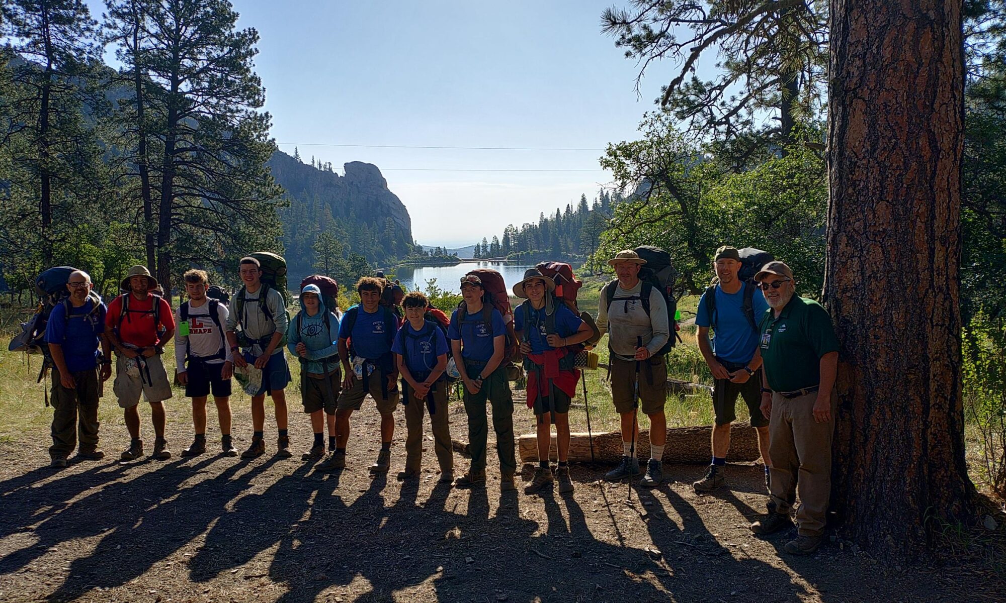 Twelve people stand next to a tree in front of a lake at the Philmont Scout Ranch in New Mexico.