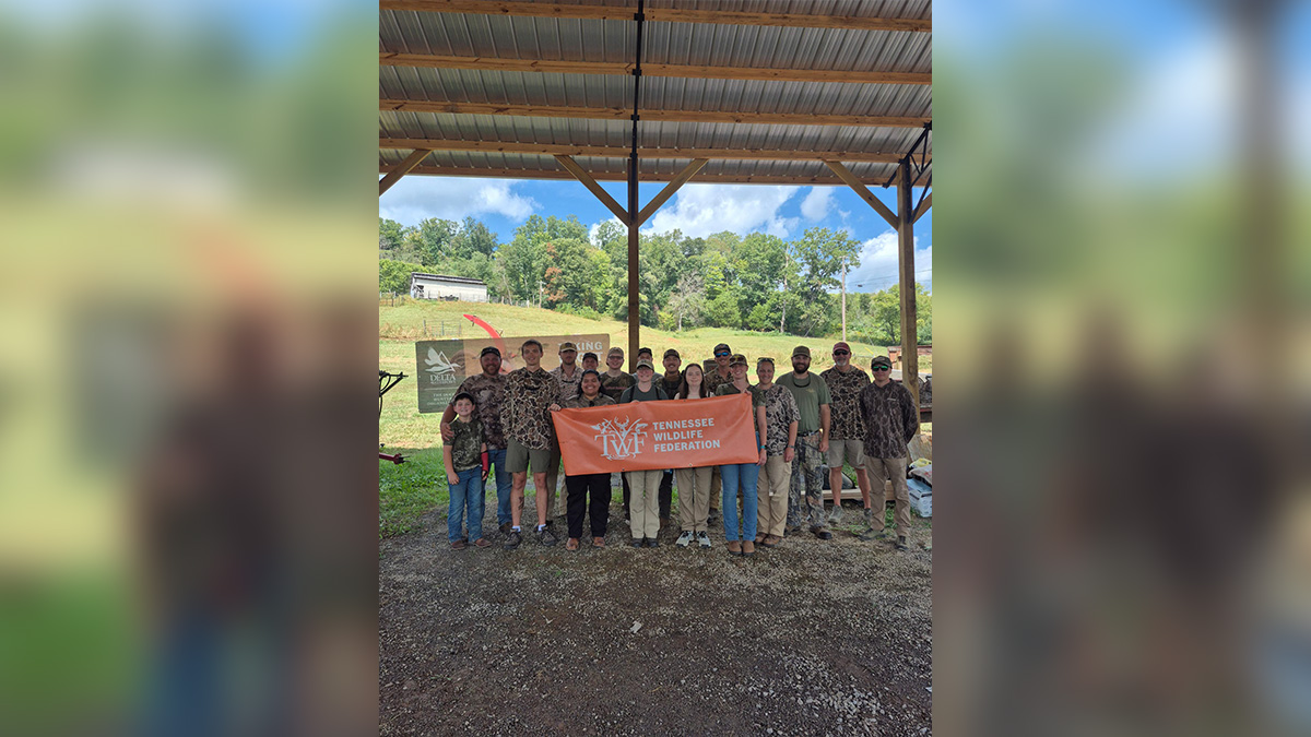 Seventeen people wearing camo stand under an awning to a barn holding an orange banner that says "Tennessee Wildlife Federation."