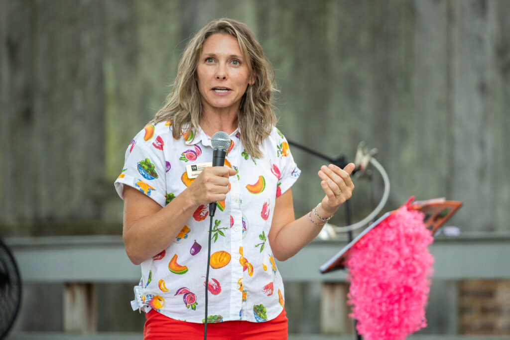 A person wearing a button up with images of fruit and red pants speaks in to a microphone.