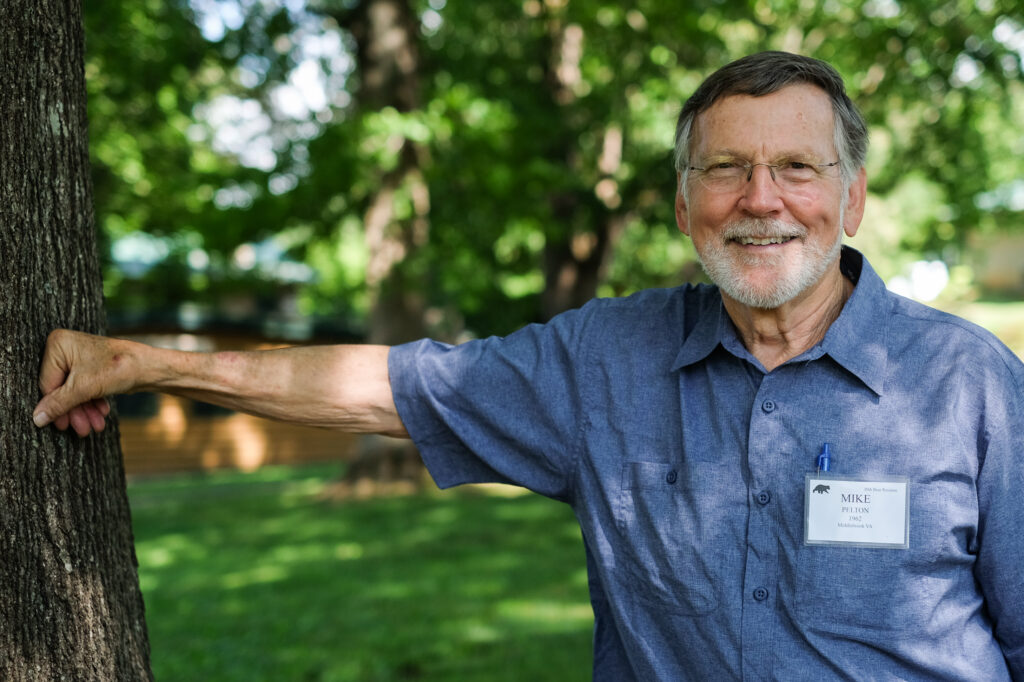 A person wearing glasses and a blue shirt leans his hand against a tree.