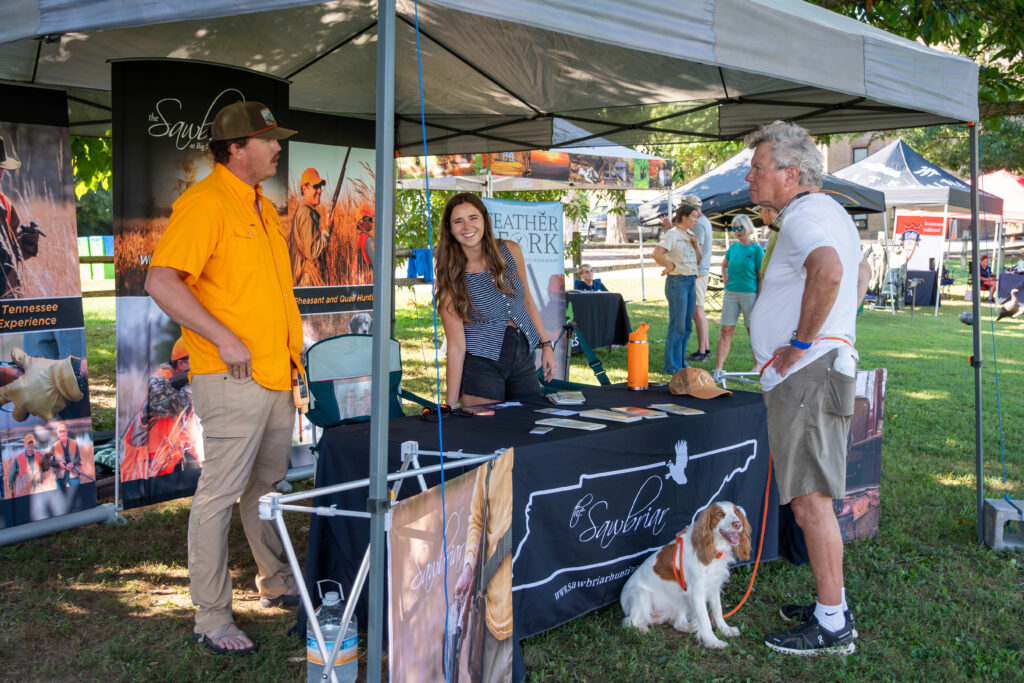 Three people stand and a dog sits near a table under a pop up tent on Maryville College campus.