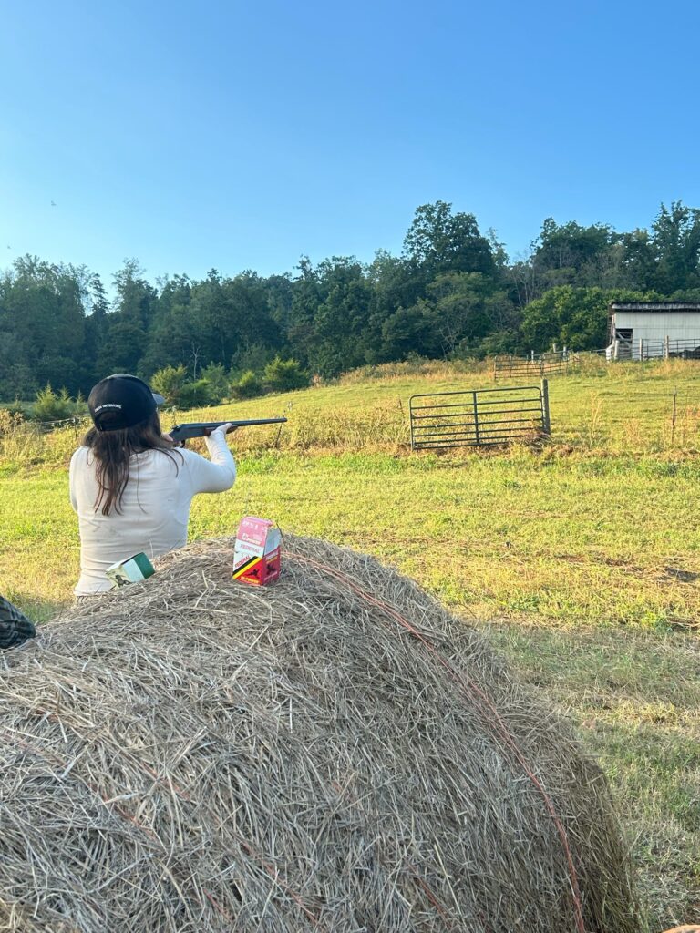 One person shoots a gun in a field near a hay bale.