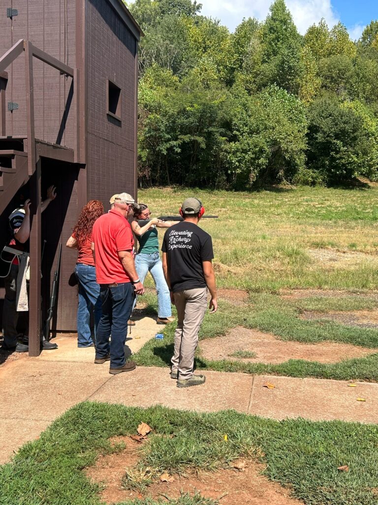 Three people instruct a fourth on shooting a gun outside a building in an empty field.