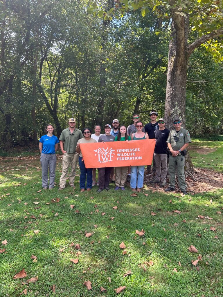 Twelve people stand in a group holding an orange banner saying "Tennessee Wildlife Federation" next to a tree.