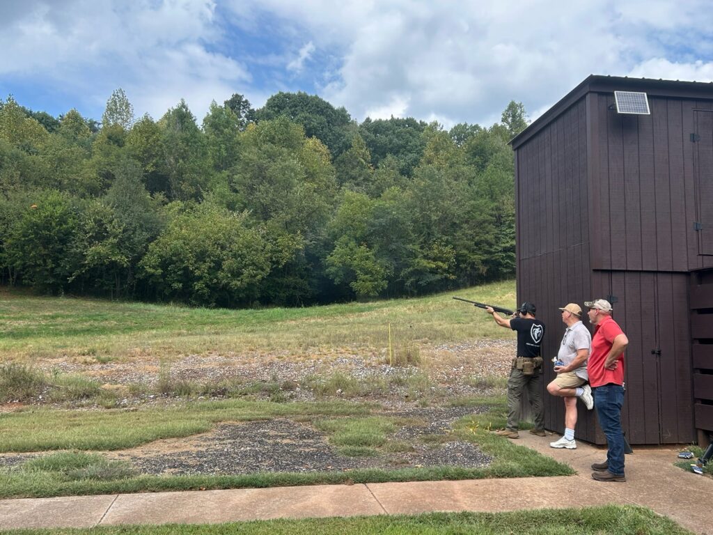 Two people stand and watch a third person shooting a gun outside a building into a field.